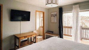 Ein Schlafzimmer mit einem Bett, einem Schreibtisch, einem Stuhl, einem Fernseher, einem Fenster mit Vorhängen und einem Balkon.