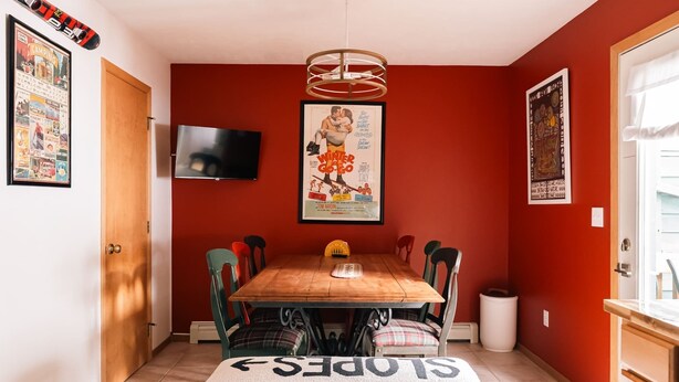 A dining area with a wooden table, chairs, a television mounted on the wall, and various posters and pictures on the walls.