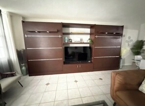 A living room with a large flat-screen TV mounted on a wooden entertainment unit, a sofa, and a coffee table. - SLEEPS 8! Beautiful & Inviting 1-BR + 1.5-BA Condo On The Beach  (Hollywood)