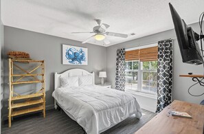 A bedroom with a bed, a ceiling fan, a window with patterned curtains, a television, and a wooden table. - A place to Wine Down in Cherry Grove! Pool, Hot Tub & Optional Golf Cart (Atlantic Beach)
