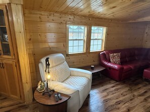 A wooden cabin interior with a white cushioned chair, a red sofa, a small table with a lamp, and a window with a view of houses.