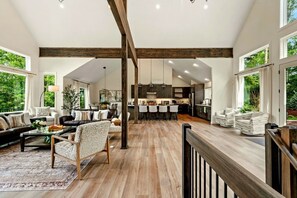 A spacious living area with a high ceiling, wooden beams, a dining area with bar stools, and large windows offering views of greenery. - Lake House Mansion | Dock+Kayaks+FirePit+Swim Spa (Long Pond)