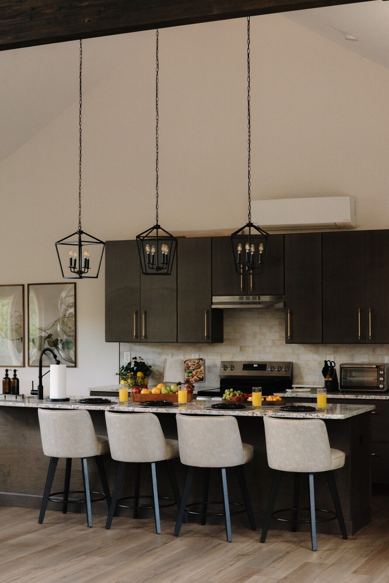 A modern kitchen with dark cabinets, a central island with bar stools, hanging pendant lights, and a stainless steel range hood.
