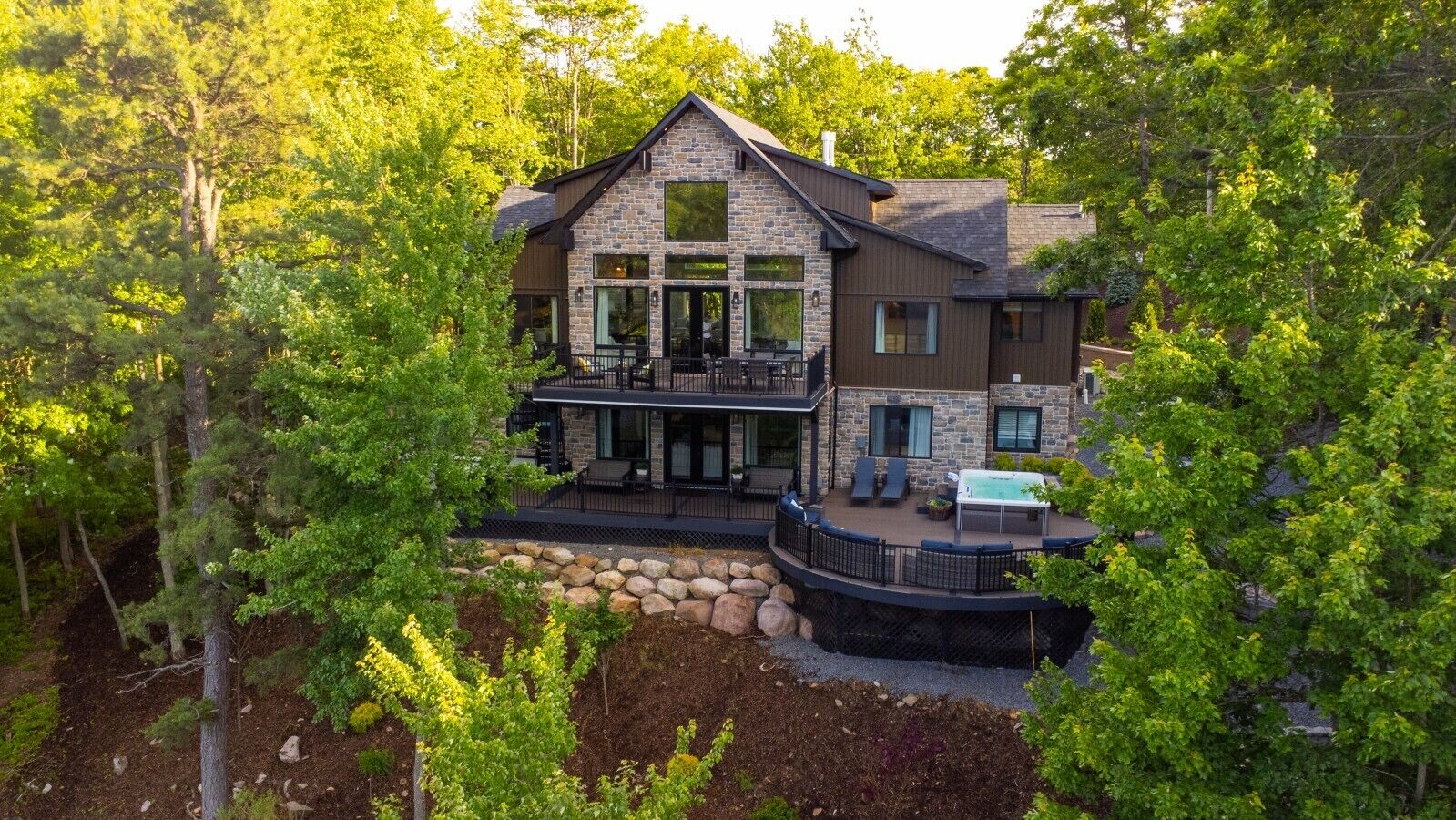 Une maison moderne à deux étages, dotée d’une façade en pierre, de grandes fenêtres et d’une terrasse aménagée avec du mobilier d’extérieur.