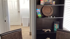 Un comedor con una mesa de vidrio, sillas de mimbre, una estantería con juegos de mesa y una cesta de objetos.