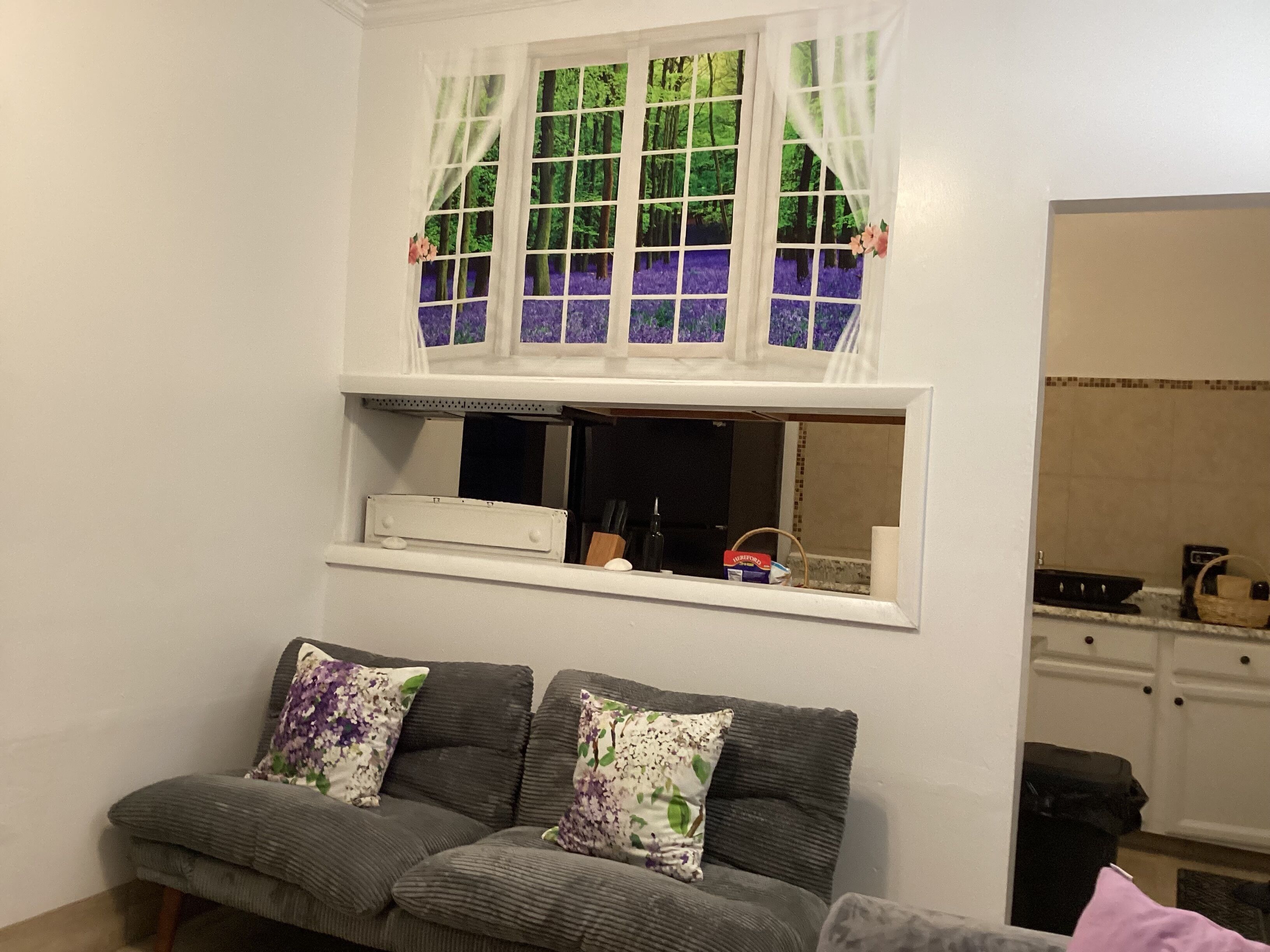 A living room with a grey sofa, a window with a view of trees, and a kitchen area in the background.