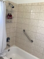 Casa de banho com banheira, chuveiro com chuveiro de mão, barra de apoio e prateleira com artigos de higiene pessoal.
