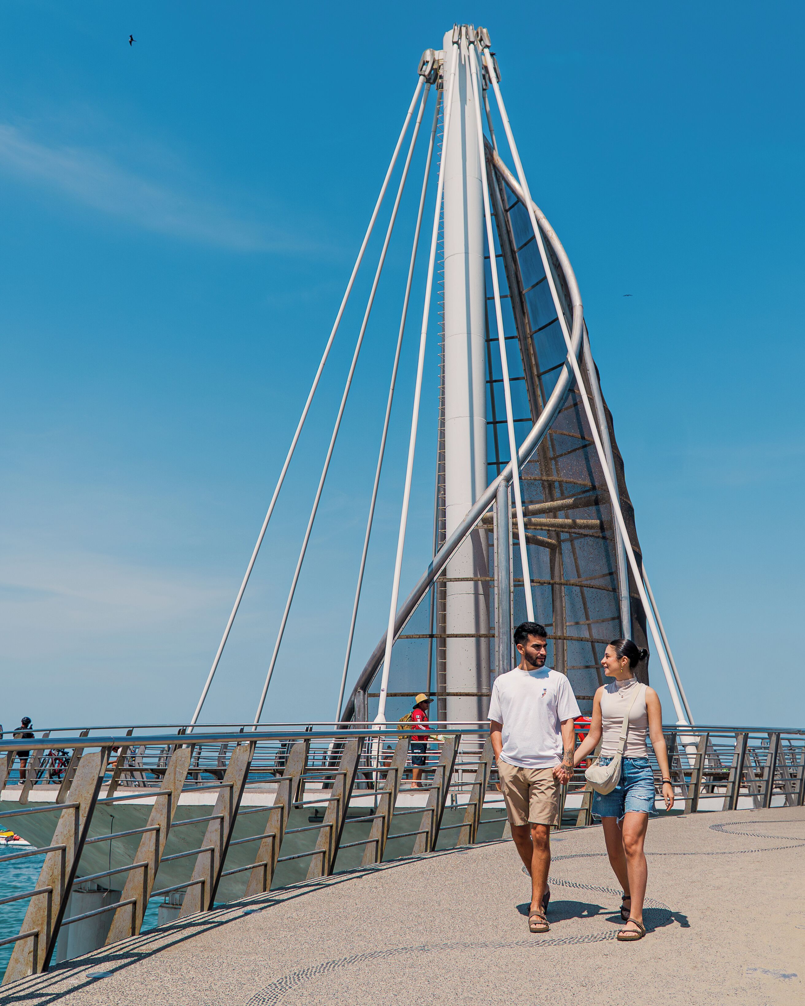 A couple walking on a pedestrian bridge with a clear sky and a tall structure in the background.