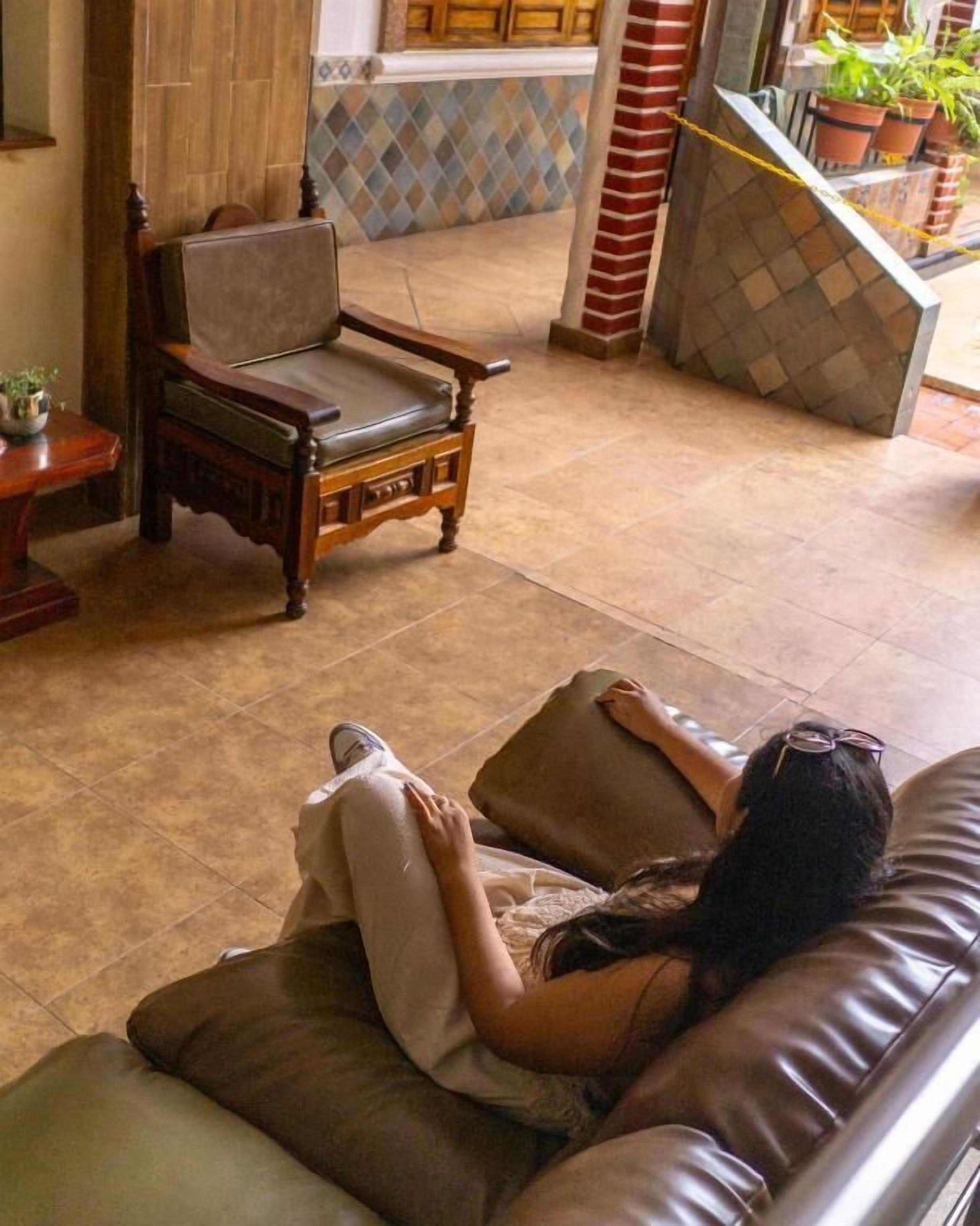 A person lying on a brown leather sofa with a cushion, a wooden chair, and a tiled floor.