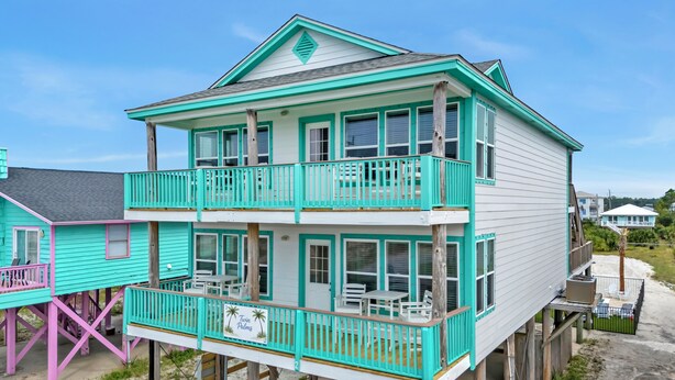 Een tweelaags huis aan het strand met een balkon, een bord met de tekst 'Twin Palms' en een zandstrand op de achtergrond.