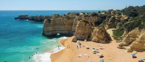 A coastal beach with clear turquoise waters, rocky cliffs, and people sunbathing on the sand.