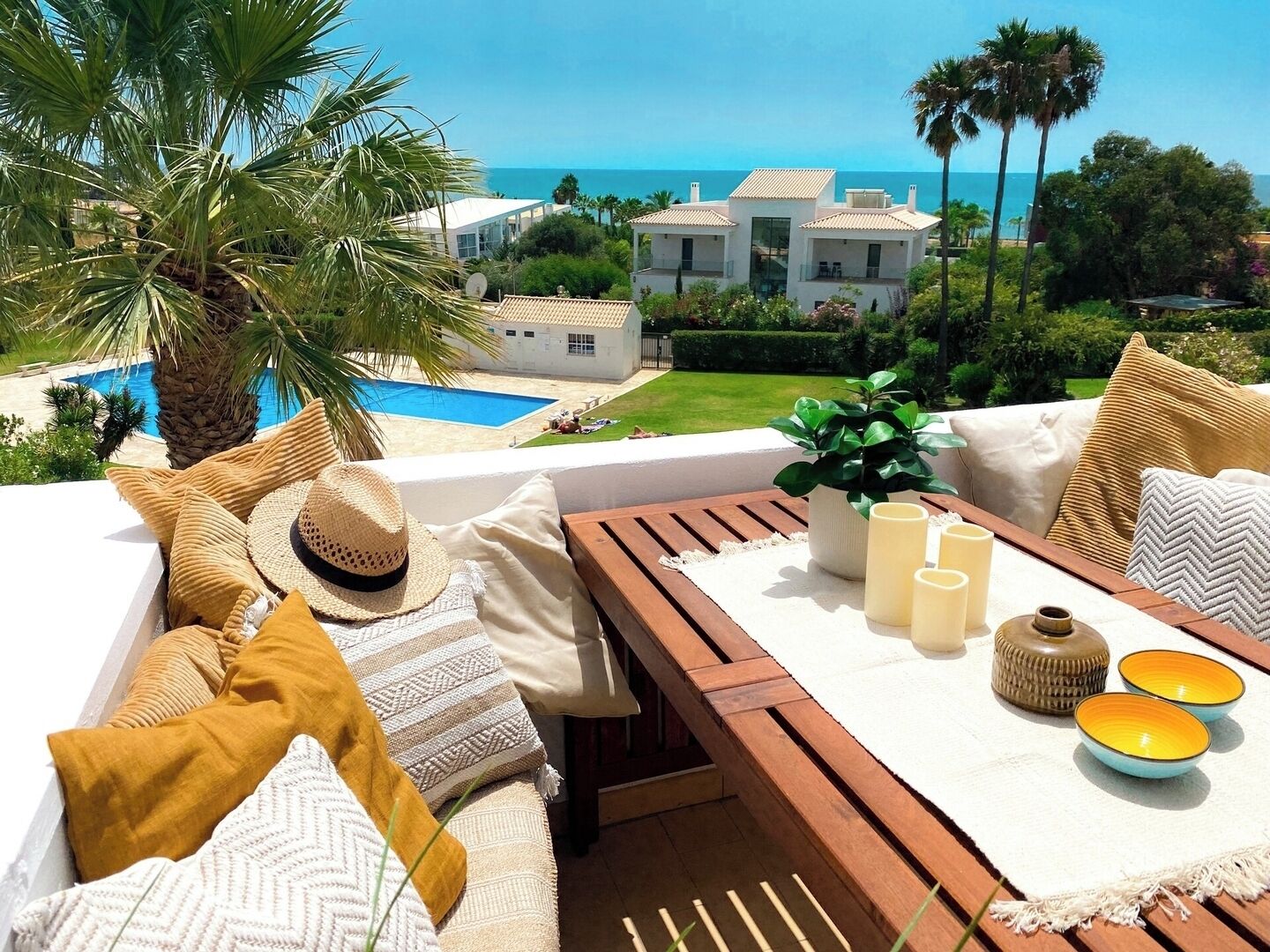 A sunlit outdoor seating area with a pool, palm trees, and a view of a coastal property.