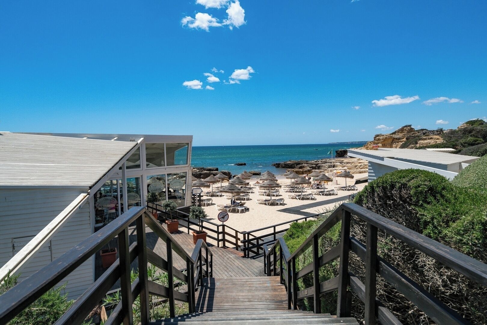 Eine Holztreppe führt zu einem Strandbereich mit Sonnenschirmen und Liegestühlen.