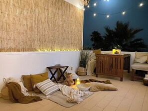 Ein Außenbereich mit Sitzgelegenheiten, einer Bambuswand, Lichterketten und einem Holztisch.