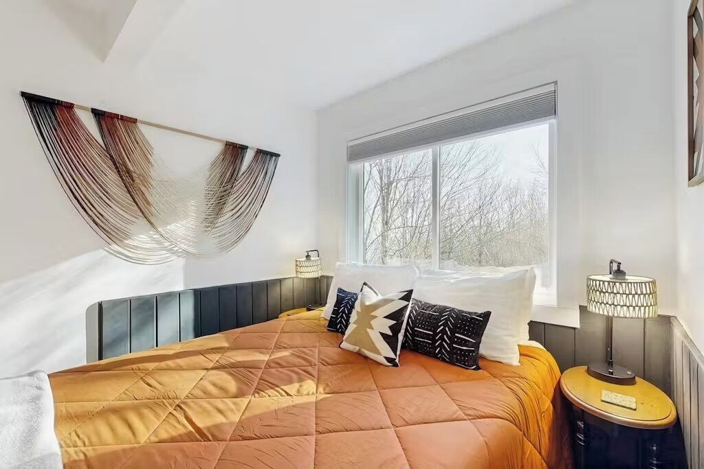 Una camera da letto moderna con un letto, cuscini, una lampada da comodino e una finestra vista alberi.