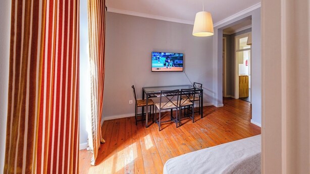 Una stanza con pavimento in legno, un tavolo da pranzo con sedie, un televisore fissato alla parete e un letto in primo piano.