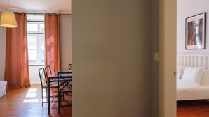 Una camera da letto con un tavolo da pranzo, sedie, una lampada a sospensione, un letto con biancheria bianca e un quadro appeso al muro.