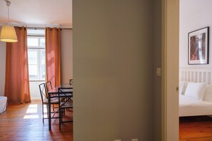 Ein Zimmer mit Esstisch, Stühlen, einer Pendelleuchte, einem Bett mit weißer Bettwäsche und einem gerahmten Bild an der Wand.