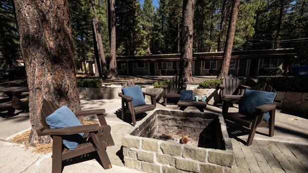 A fire pit surrounded by wooden chairs and benches, set in a wooded area with trees and a building in the background. - Charming 2 Queen bedroom lodge in South Lake Tahoe for a relaxing stay (South Lake Tahoe)