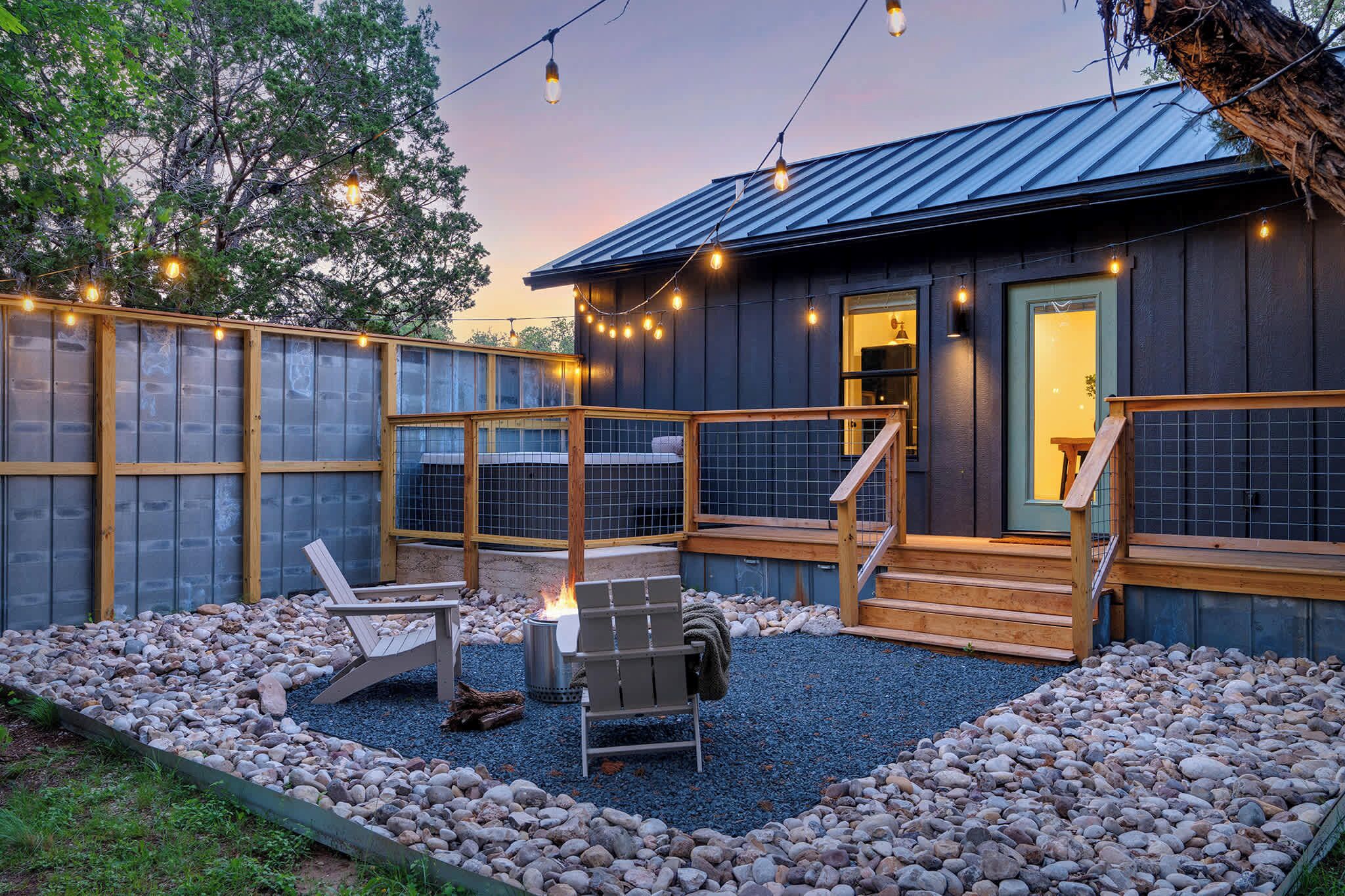A wooden cabin with a deck, two Adirondack chairs, a fire pit, and string lights.