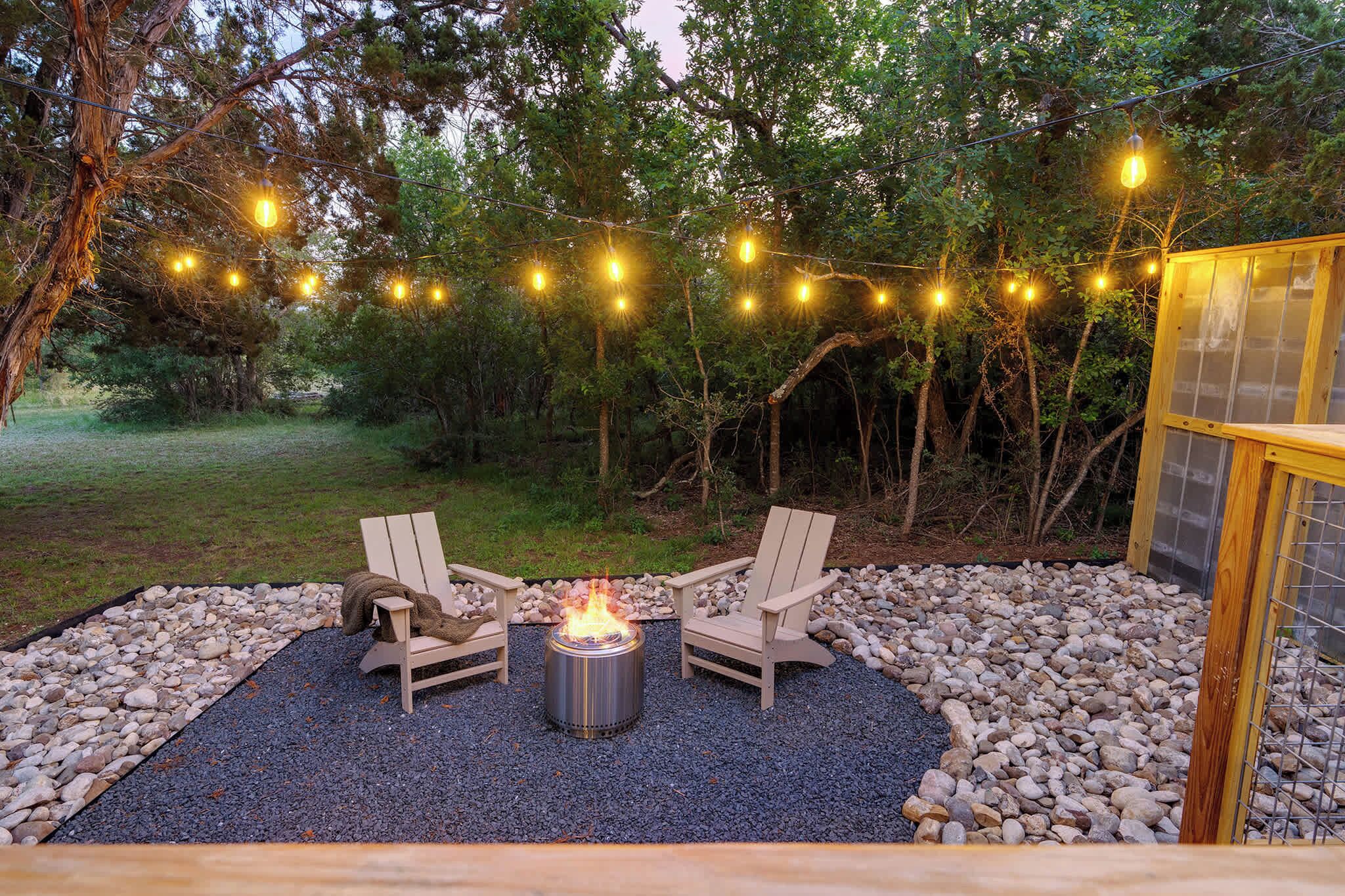 An outdoor seating area with two Adirondack chairs around a fire pit, surrounded by a gravel and pebble border, with string lights overhead.