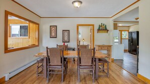 Un coin repas avec une table et des chaises en bois, un luminaire au plafond, des tableaux encadrés au mur et une vue sur la cuisine.