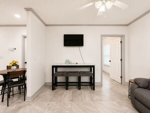 A modern living room with a dining area, a flat-screen TV mounted on the wall, and a ceiling fan.