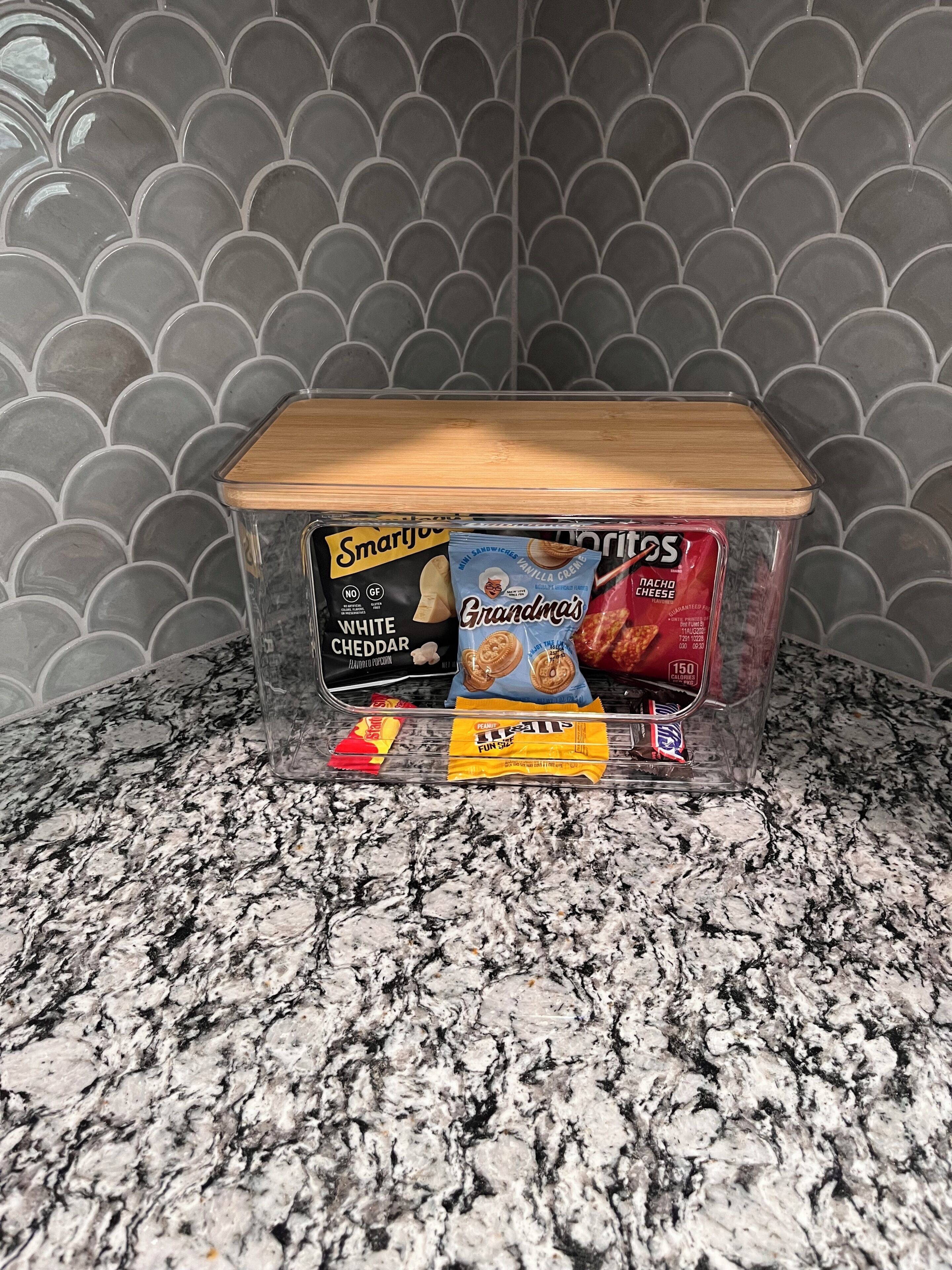 A clear plastic storage container with a wooden lid on a granite countertop. Inside, there are several snack items including SmartJoy white cheddar popcorn, Grandma's vanilla creme cookies, and a bag of nacho cheese.