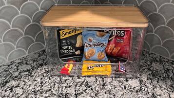 A clear plastic storage container with a wooden lid on a granite countertop. Inside, there are several snack items including SmartJoy white cheddar popcorn, Grandma's vanilla creme cookies, and a bag of nacho cheese.