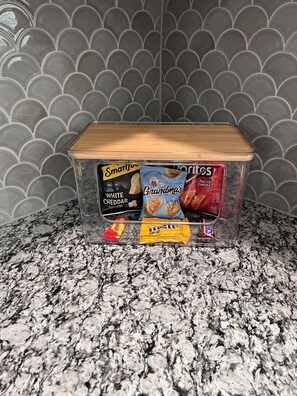 Un récipient de rangement transparent en plastique, doté d’un couvercle en bois, est posé sur un comptoir en granit. À l’intérieur, on trouve divers en-cas, notamment du pop-corn au fromage blanc de la marque SmartJoy, des biscuits à la crème vanille de la marque Grandma’s et un sachet de fromage pour nachos.