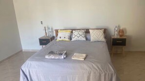 A neatly made bed with a grey bedspread, two bedside tables with water bottles, and a small table with a glass. - Bright and Cozy Apartment in a Central Location (Béjaïa)