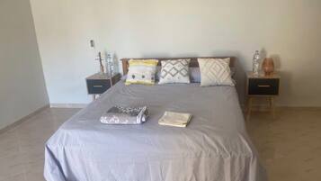 A neatly made bed with a grey bedspread, two bedside tables with water bottles, and a small table with a glass.