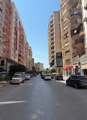 A street lined with multi-story residential buildings, parked cars, and a clear sky. - Bright and Cozy Apartment in a Central Location (Béjaïa)