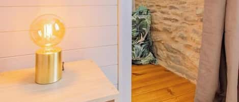 A room with a wooden floor, a small table with a lamp, a stone wall, and a window with curtains.