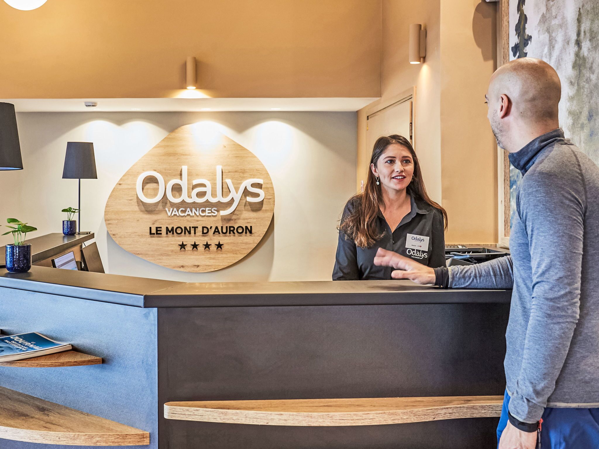 A reception desk with a wooden sign that reads 'Odalys Vacances Le Mont d'Auron' and a name tag for a staff member.