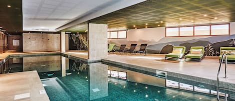 An indoor swimming pool with a tiled floor, lounge chairs, and a wall mural.