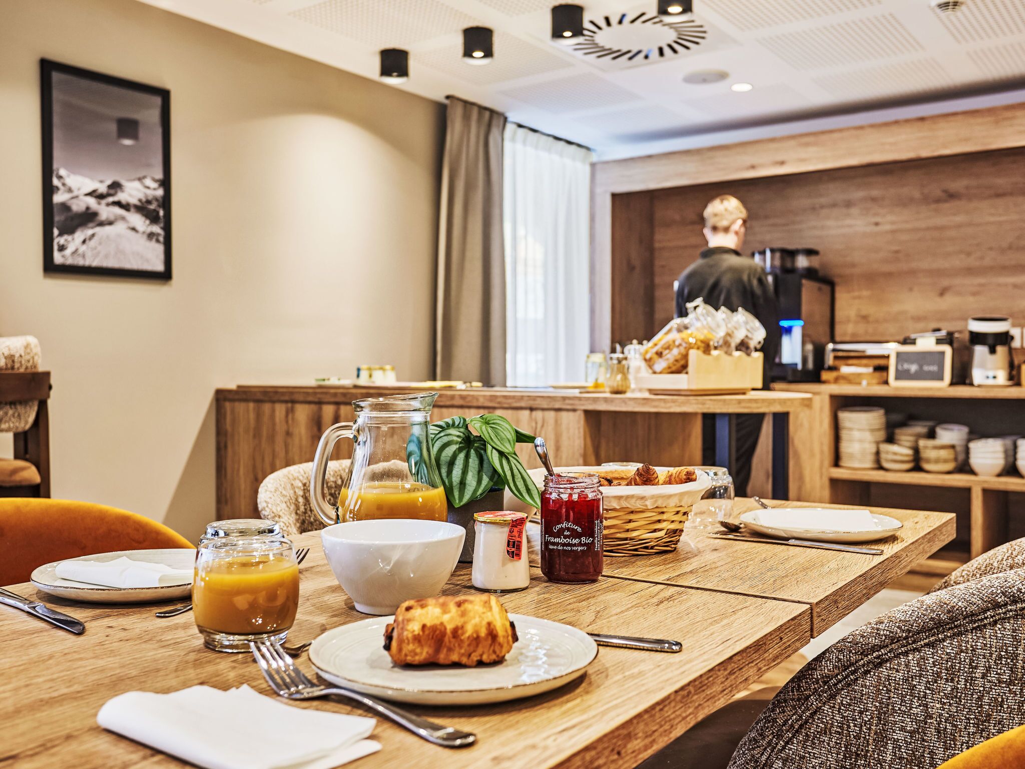 A breakfast setting with a table set for two, including a croissant, a glass of orange juice, a bowl, and a jar of jam.