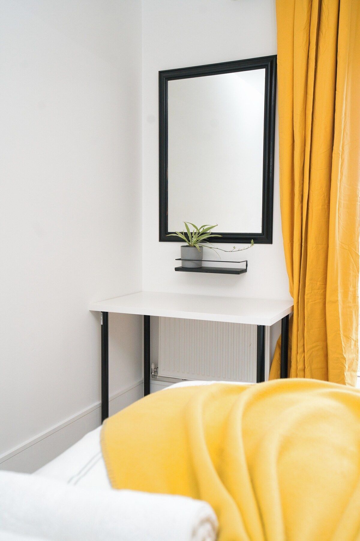A hotel room with a white bed, a bedside table with a potted plant, a black-framed mirror, and yellow curtains.
