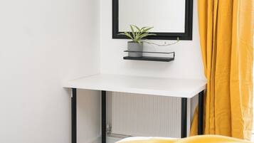A hotel room with a white bed, a bedside table with a potted plant, a black-framed mirror, and yellow curtains.