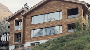 A modern two-story building with large windows and a wooden facade, set against a mountainous backdrop.