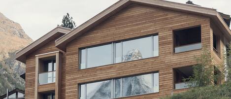 A modern two-story building with large windows and a wooden facade, set against a mountainous backdrop.