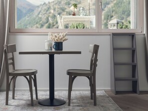 Ein Zimmer mit Tisch und zwei Stühlen, ein Fenster mit Bergblick, eine Leiter und ein kleiner Tisch mit Vase und Pflanze.