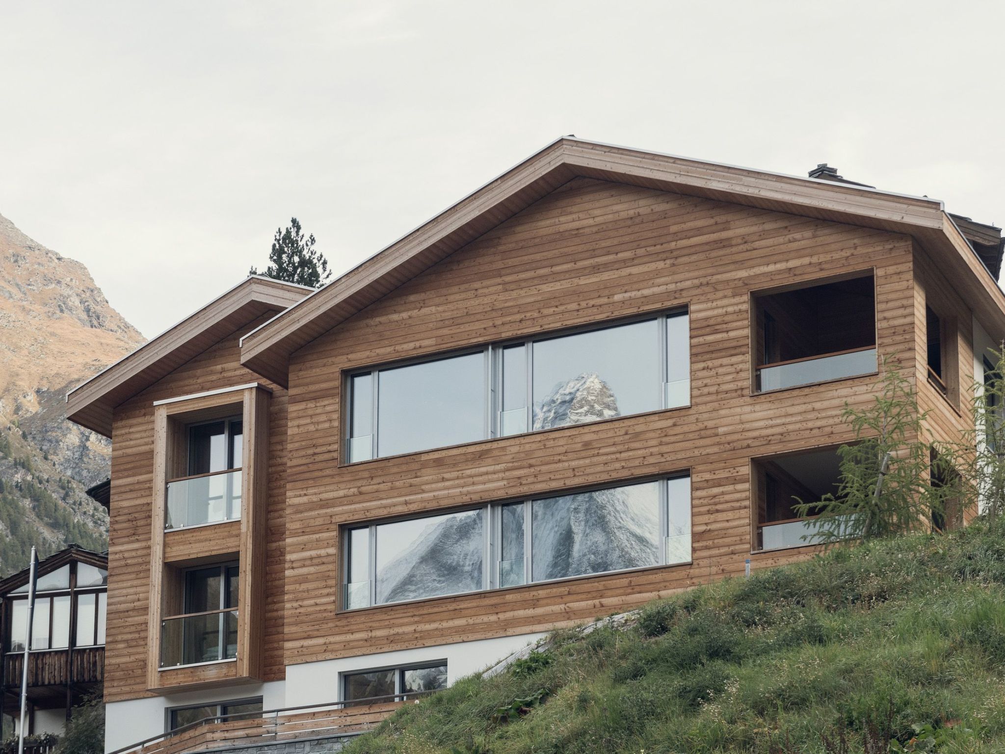 Un moderno edificio de varios pisos con grandes ventanales y fachada de madera, con montañas al fondo.