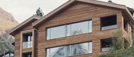A modern multi-story building with large windows and a wooden facade, set against a mountainous backdrop.