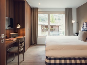 A hotel room with a bed, a desk with a chair, a television, and a view of a building through a window. - OVERLOOK Lodge by CERVO Zermatt, (Zermatt), by Interhome (Zermatt)