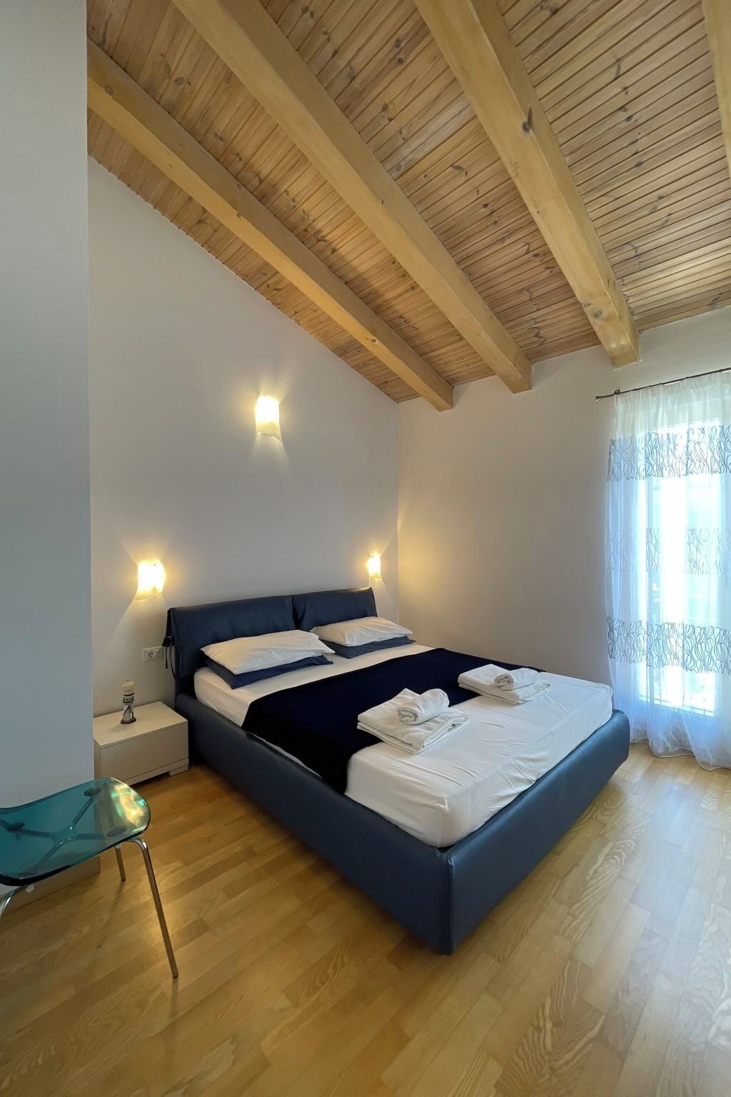 Une chambre à coucher avec un plafond en bois, un lit recouvert de draps blancs, deux lampes de chevet et une fenêtre ornée de rideaux légers.
