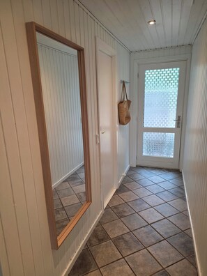 Ein Flur mit weiß gepanelten Wänden, einem spiegelnden Rahmen aus Holz, einer Tür mit Milchglasfüllung und einem gefliesten Boden.