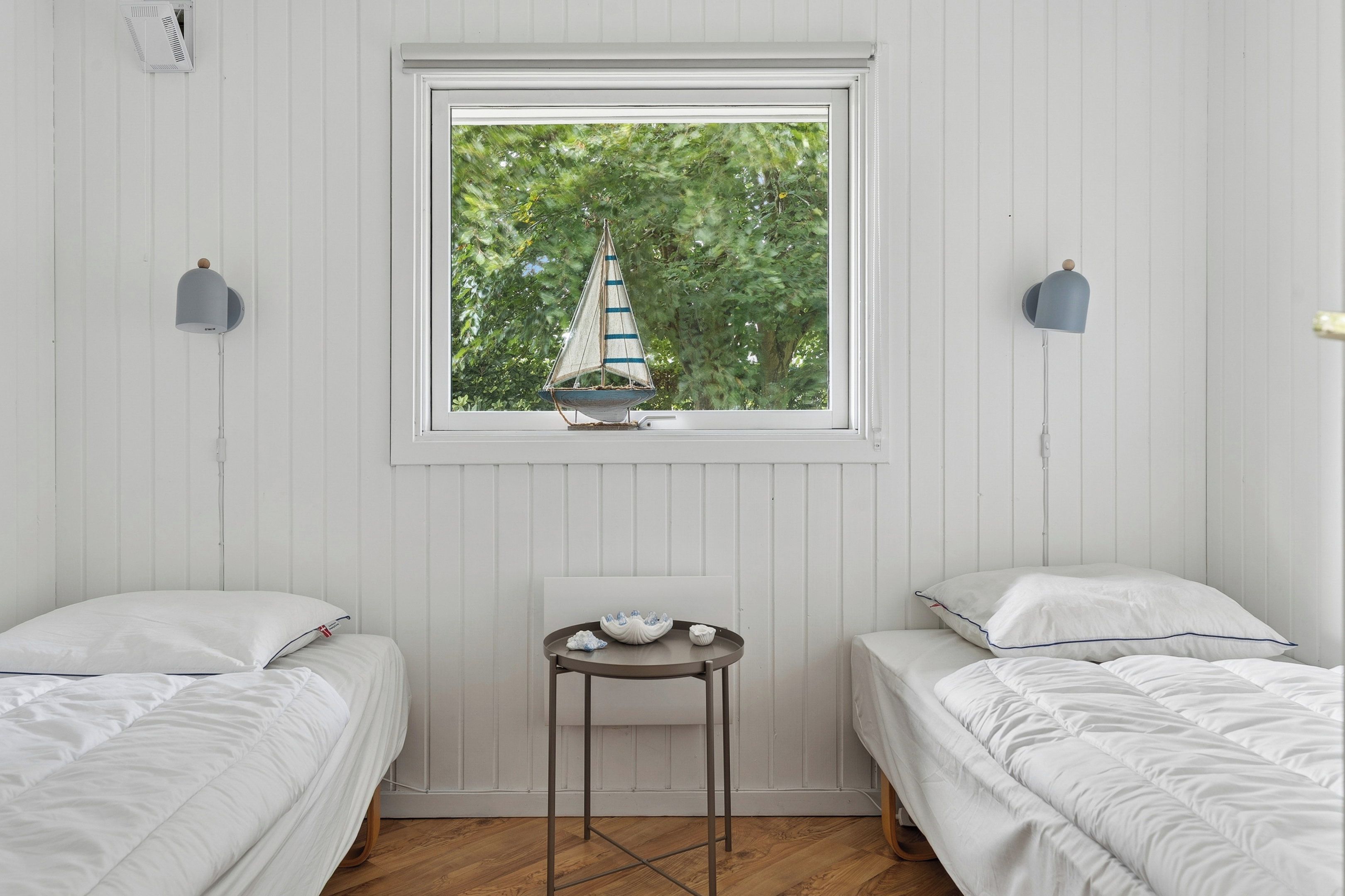 A bedroom with two beds, a small table, and a window with a sailboat decoration.