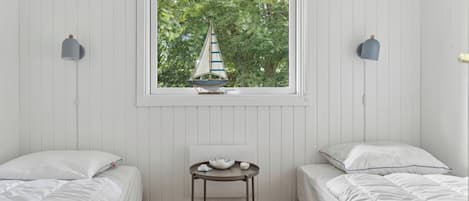A bedroom with two beds, a small table, and a window with a sailboat decoration.