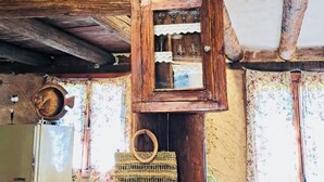 A rustic interior with a wooden ceiling, a small window with lace curtains, and a woven basket hanging on the wall.